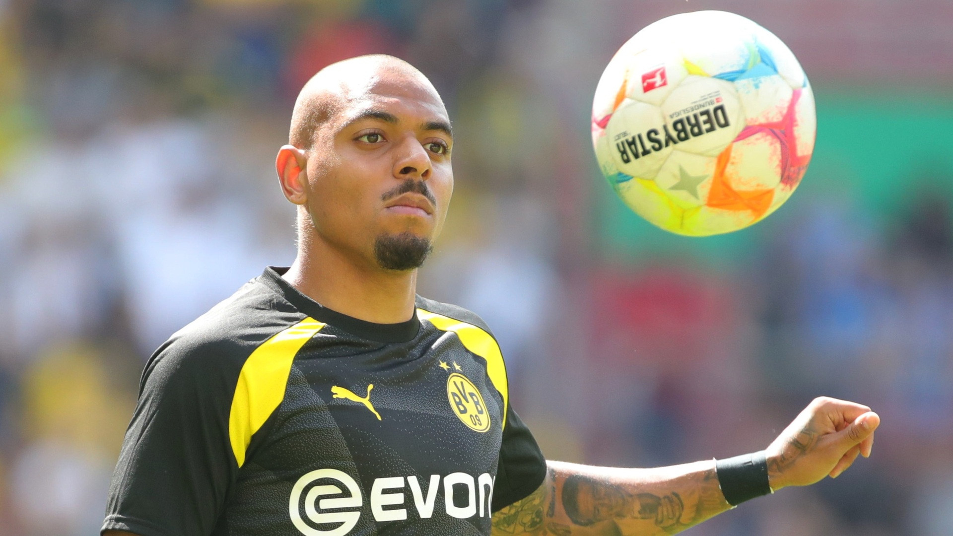 Action photograph of Donyell Malen in black and yellow kit as a football floats in midair beside his head.