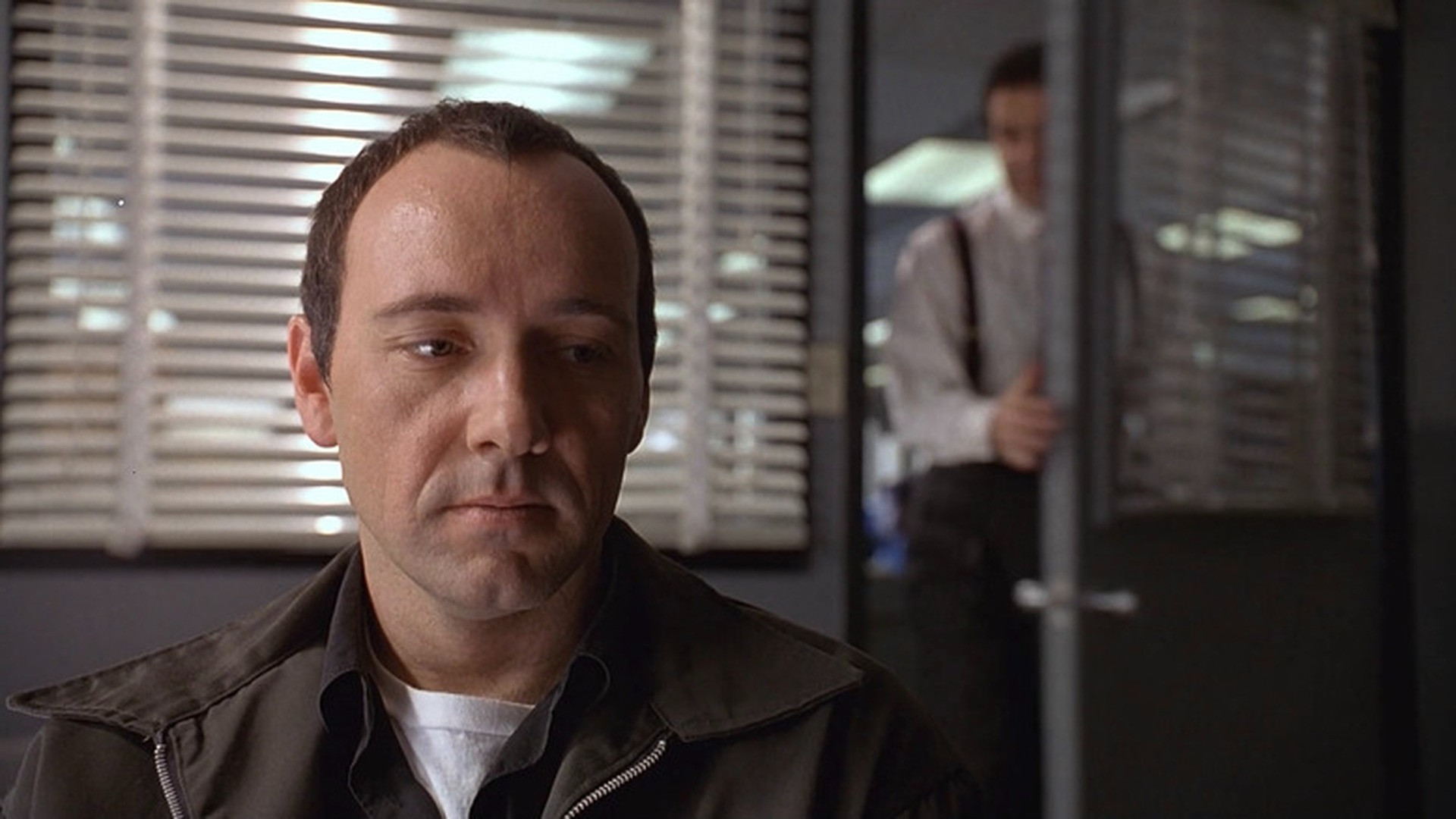 Verbal Kint looking downward with a pensive expression during police interrogation, image horizontally flipped.