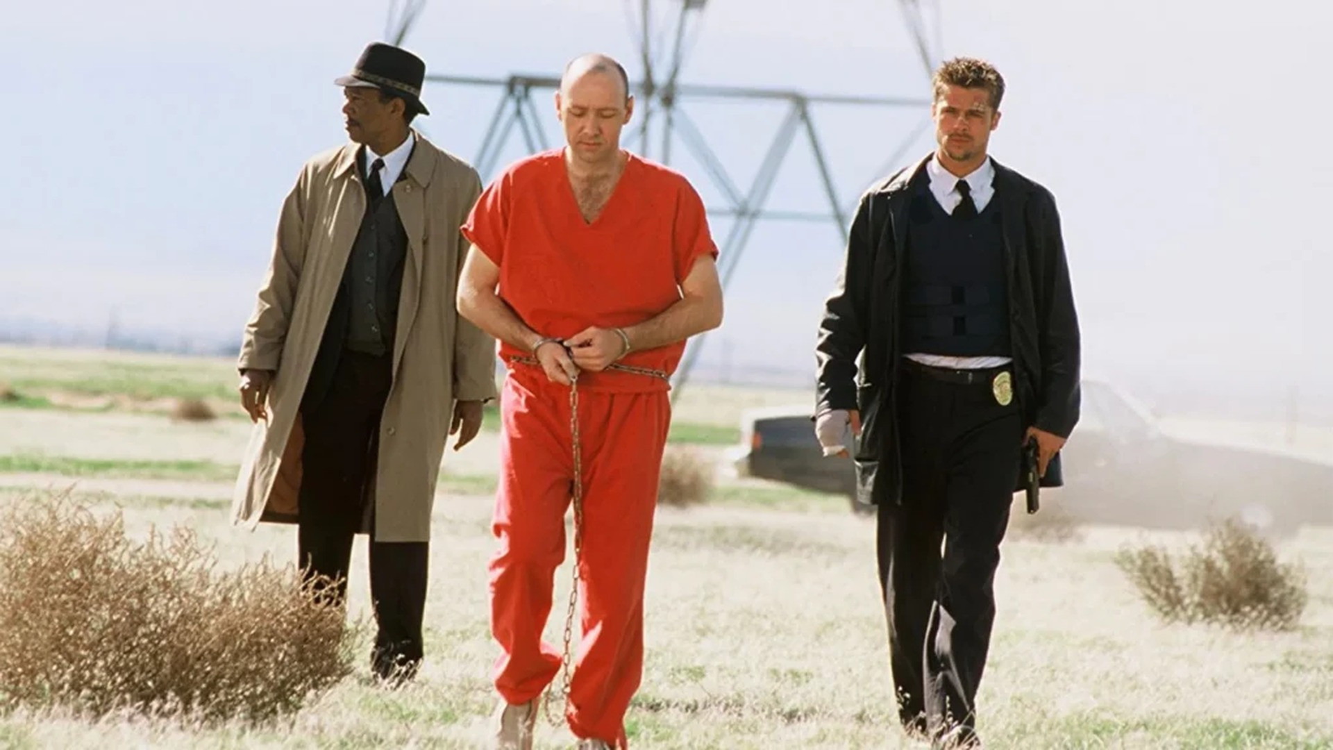Detectives Mills and Somerset escorting handcuffed suspect John Doe in an orange jumpsuit across an open field, image horizontally flipped.