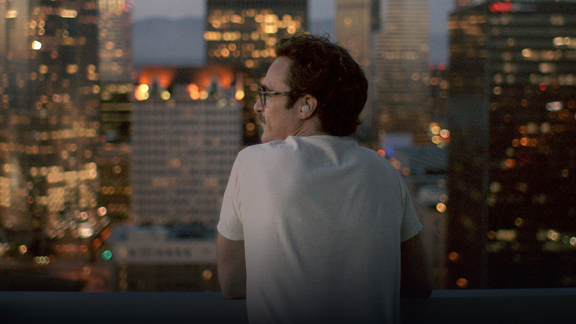 Joaquin Phoenix in Her walking through a sunlit city speaking into a discreet wireless earpiece.